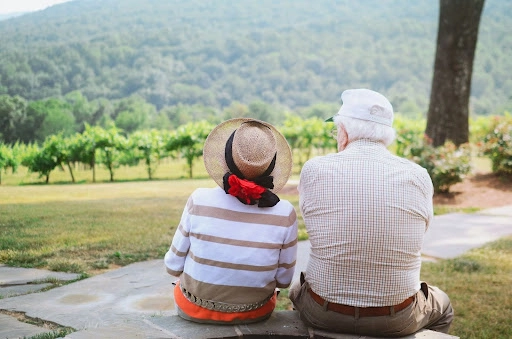 An elderly couple sitting outdoors on a stone pathway, overlooking a scenic landscape with lush greenery and rolling hills.